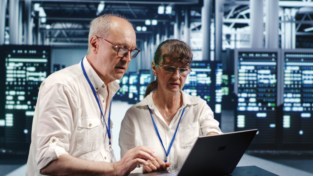 Two adults in white shirts, wearing lanyards with badges, focus intently on a laptop in a IT Security server room. The background shows blurred computer servers.