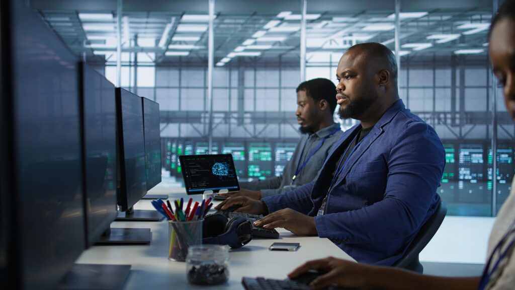 A group of focused IT Security professionals work at desks with multiple monitors displaying data in a modern, high-tech control room setting.