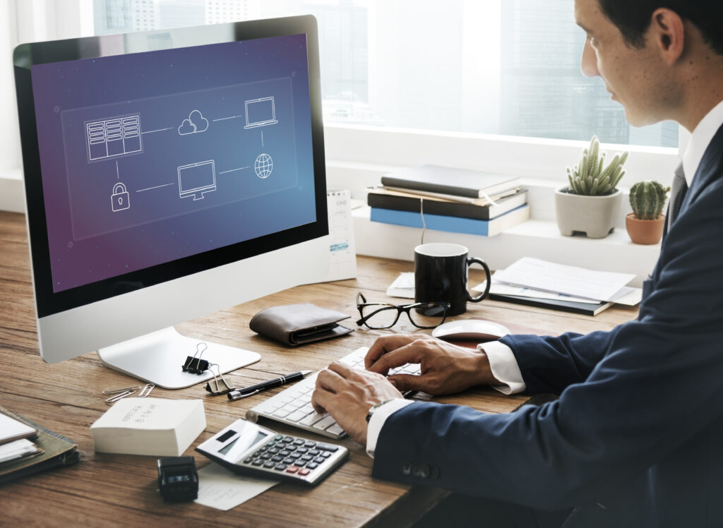 A man in a suit works at a desk with a computer displaying a network diagram. The office setting is organized, conveying a focused, professional tone.