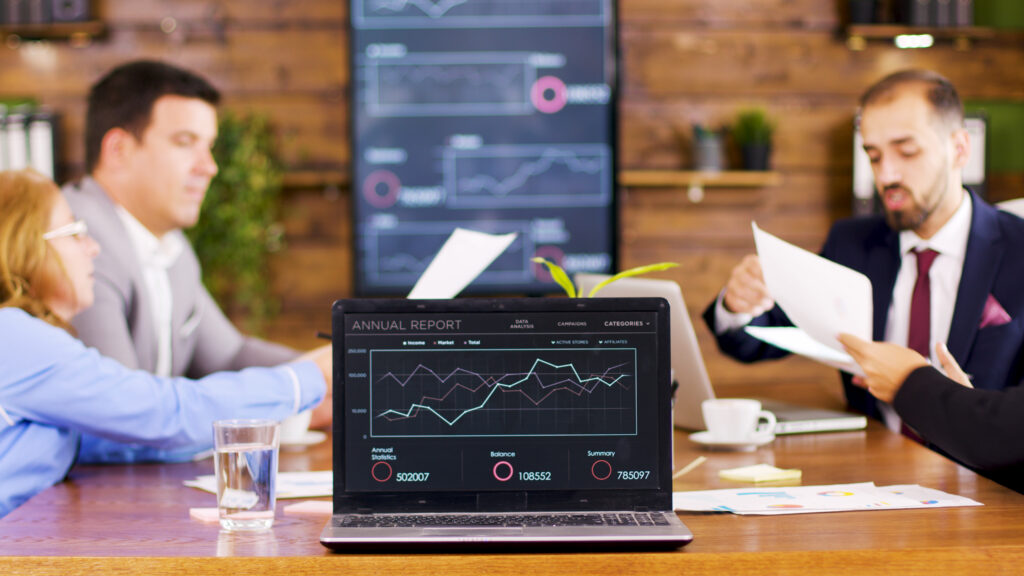 A group of professionals engaged in a meeting around a table, with a laptop displaying an annual report chart. The atmosphere is focused and collaborative.