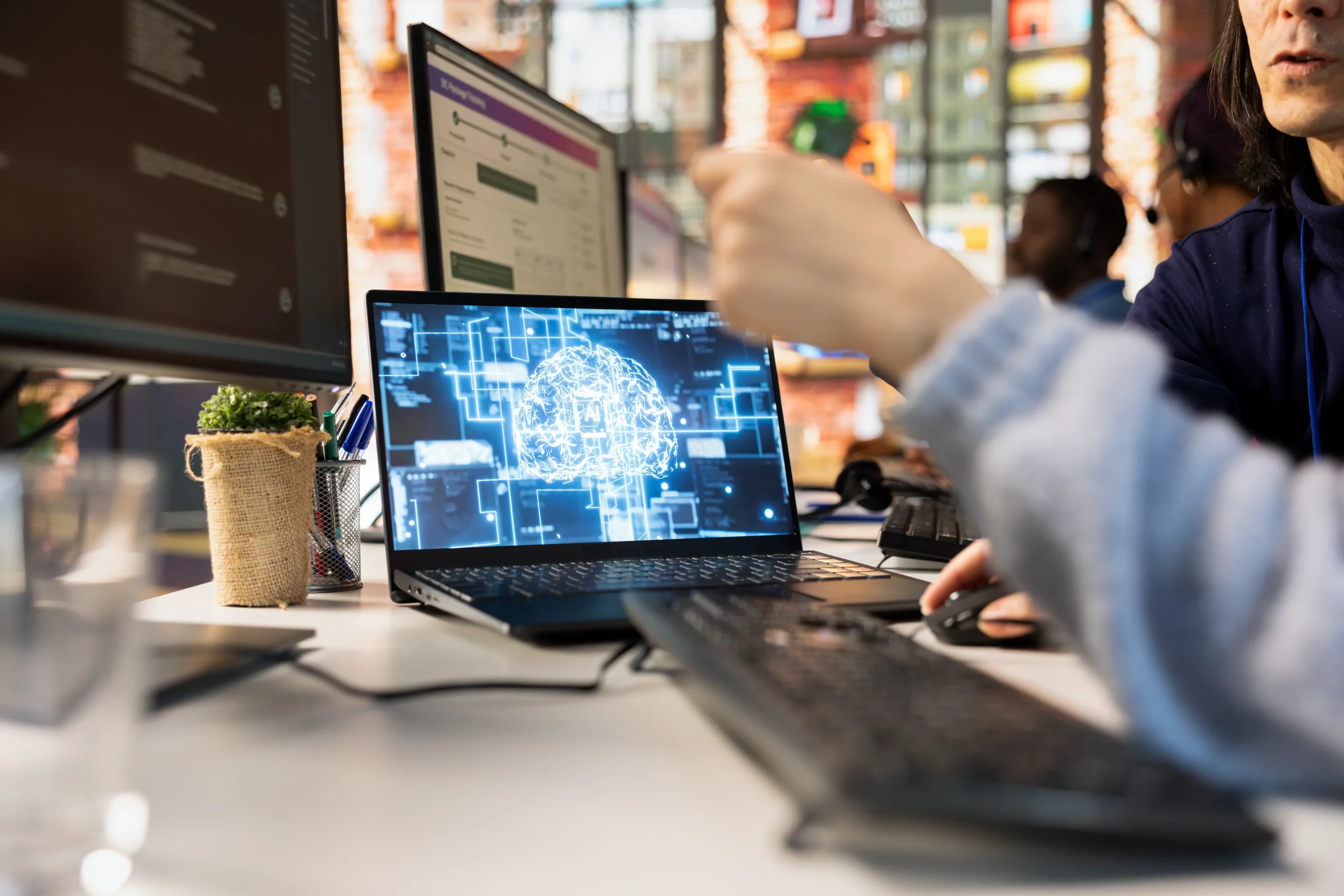 A person points to a laptop screen displaying a digital brain graphic, surrounded by monitors in a tech-filled office. The atmosphere is collaborative and focused Business technology services netsectechnologies