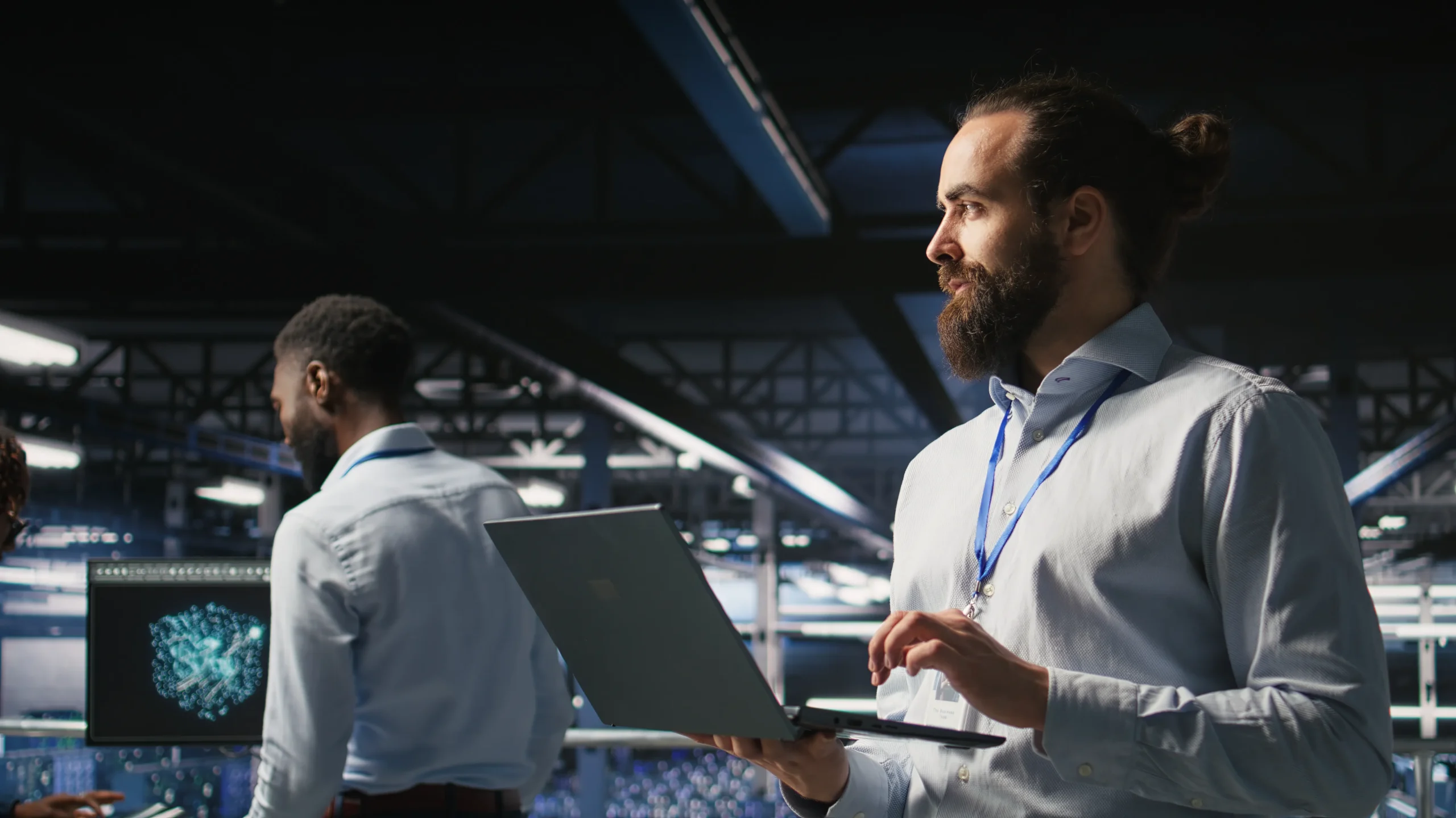 A man with a beard holds a laptop in a modern, brightly lit tech environment. Another person examines data on a wall screen. The scene is focused and professional Business technology services netsectechnologies