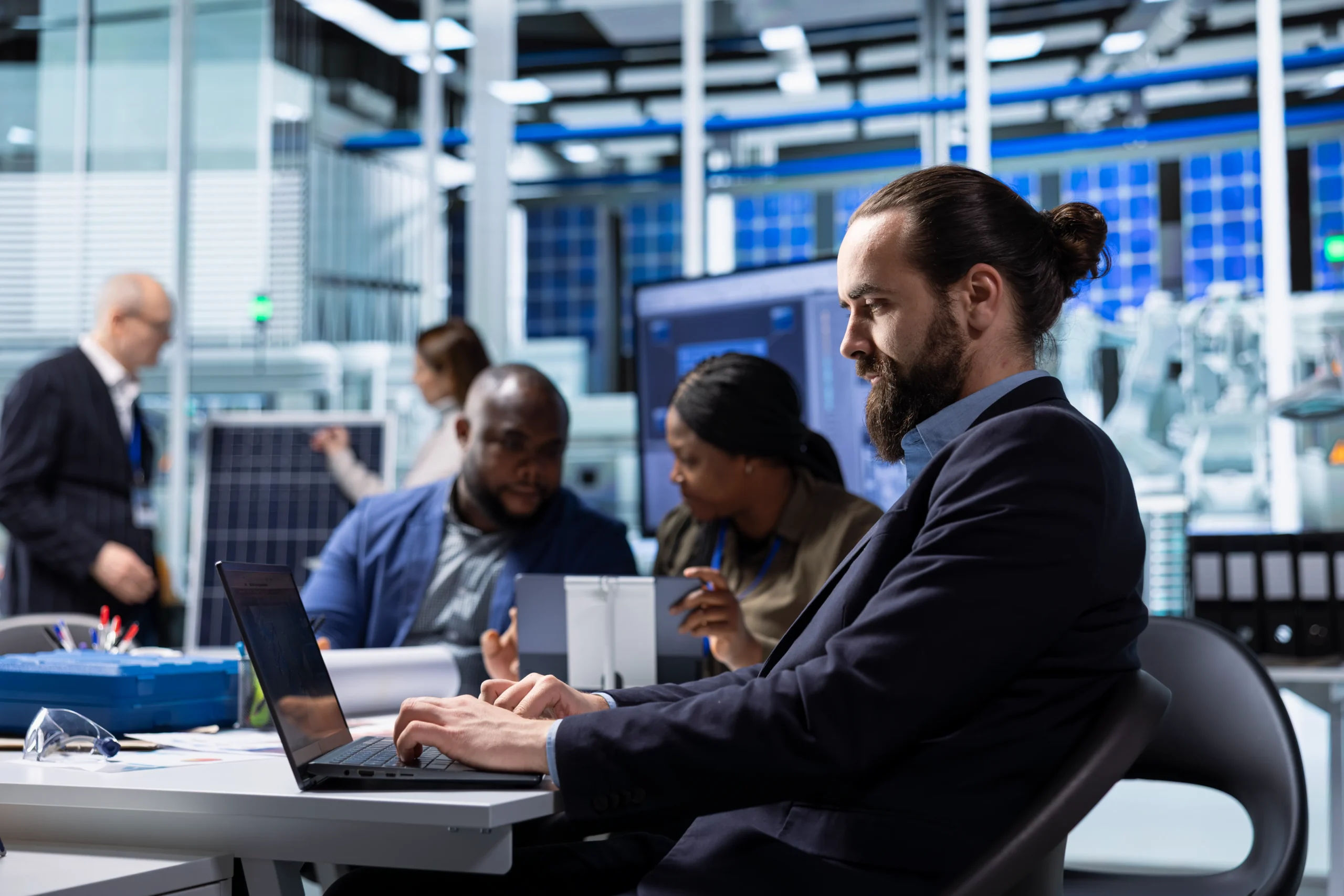 Man with a beard and bun types on a laptop at a conference table in a tech lab. Colleagues in background discuss and examine equipment Business technology services netsectechnologies