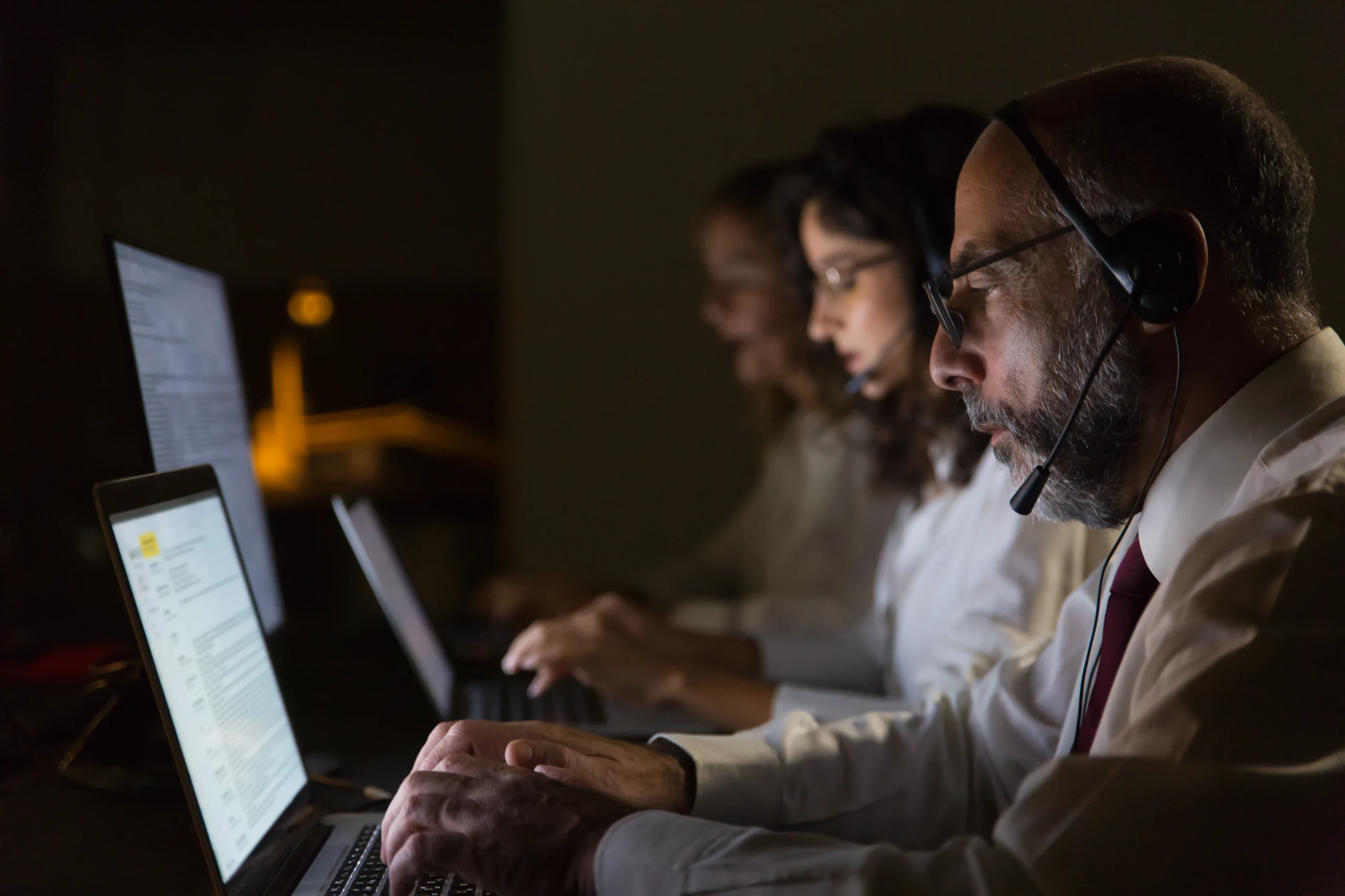 Three focused individuals sit in a dimly lit room working on laptops. The person in the foreground wears a headset, suggesting a call center environment 24/7 IT support at netsectechnologies