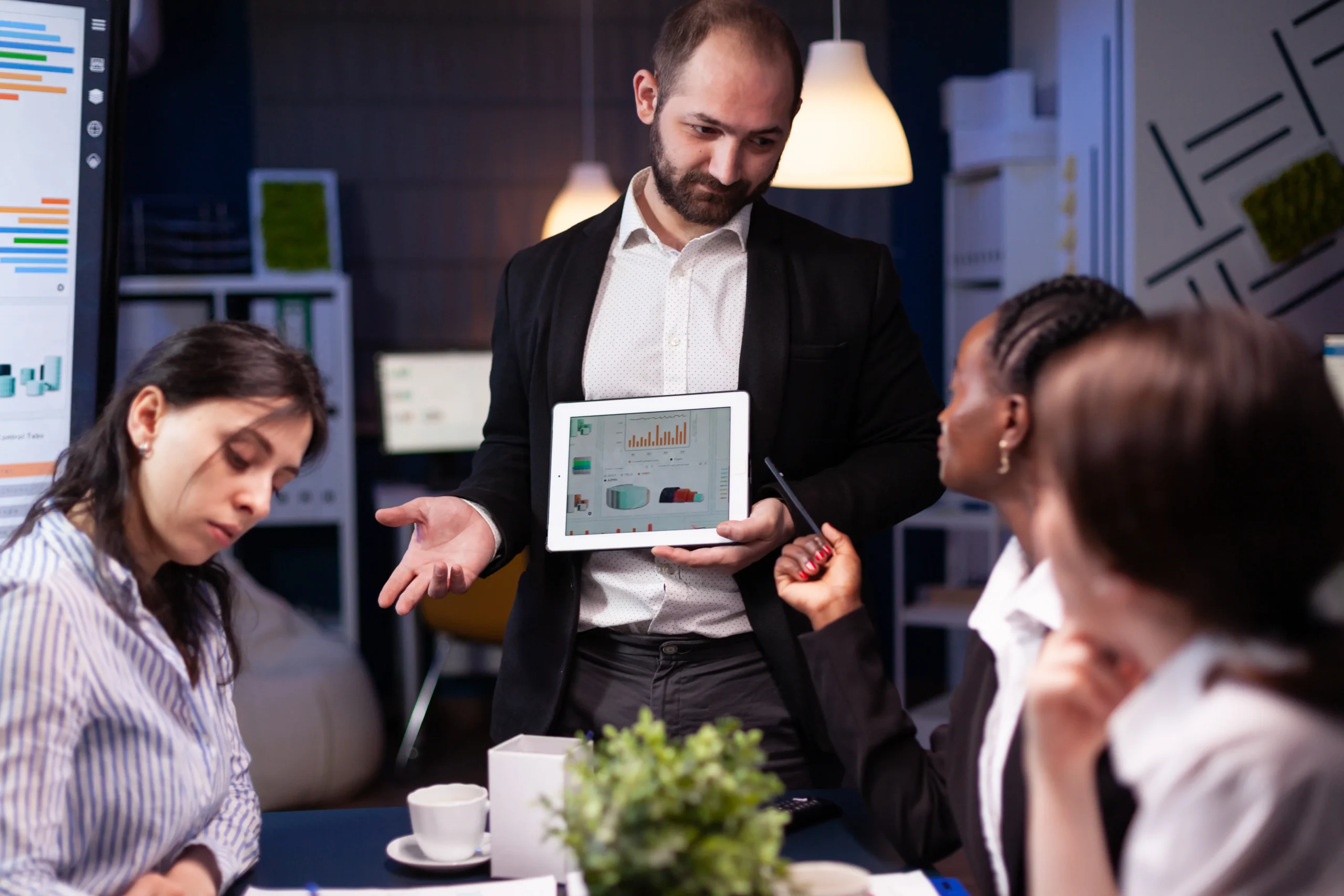 A man in business attire presents data on a tablet to three colleagues in a modern office. Charts are visible on screens; a focused discussion ensues Business technology services netsectechnologies