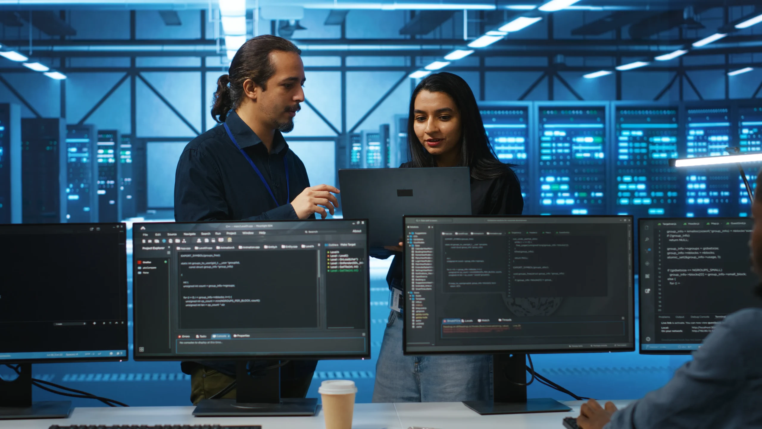 Two people discuss over a laptop in a server room with large screens displaying code. The setting is high-tech and collaborative managed it services with netsectechnologies