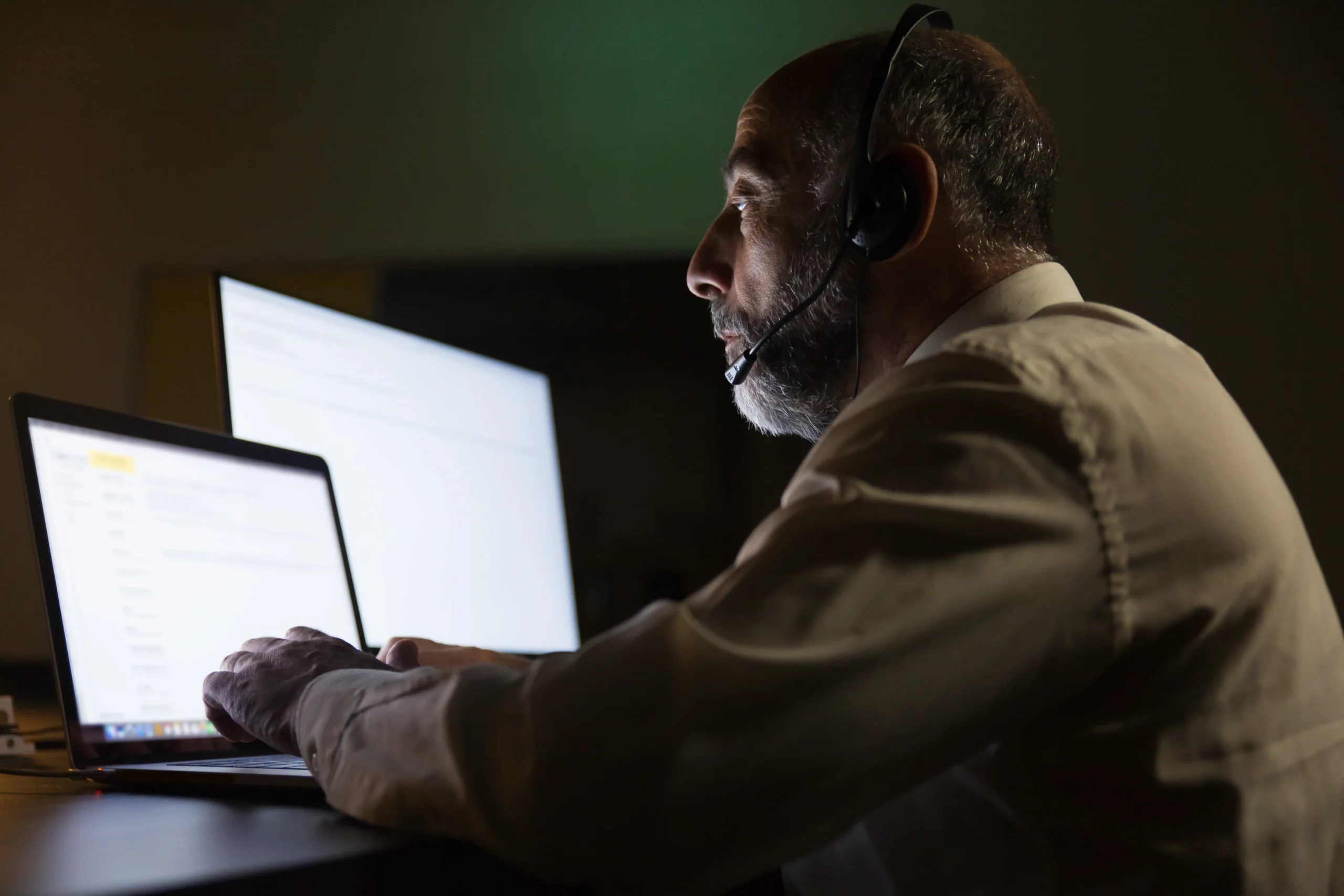 A man wearing a headset sits at a desk in a dimly lit room, focused on a laptop and a bright monitor. The scene conveys concentration and focus 24/7 IT support at netsectechnologies