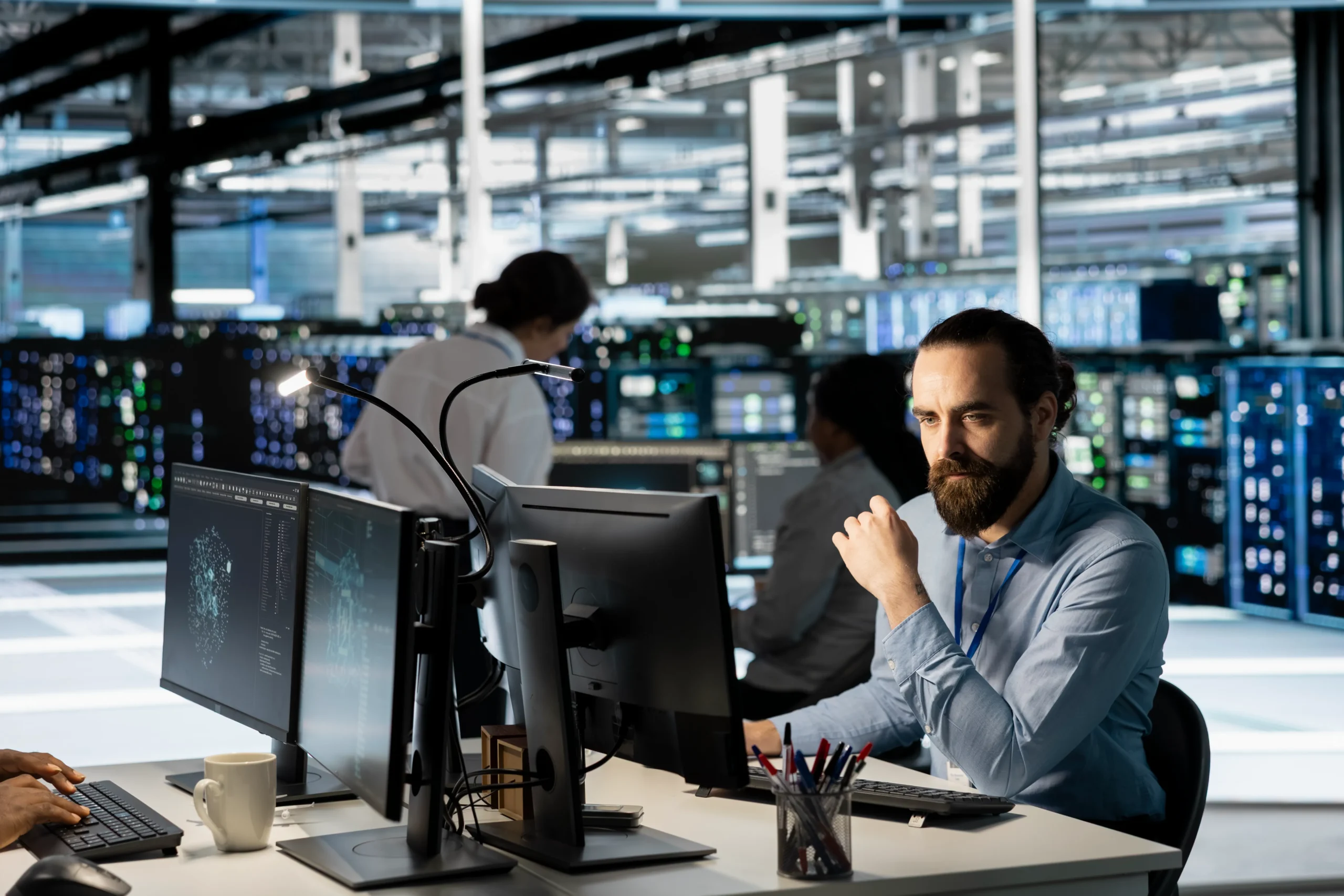 A man with a beard focused on computer screens in a high-tech server room. Colleagues work in the background. The atmosphere is professional and intense 24/7 IT support at netsectechnologies