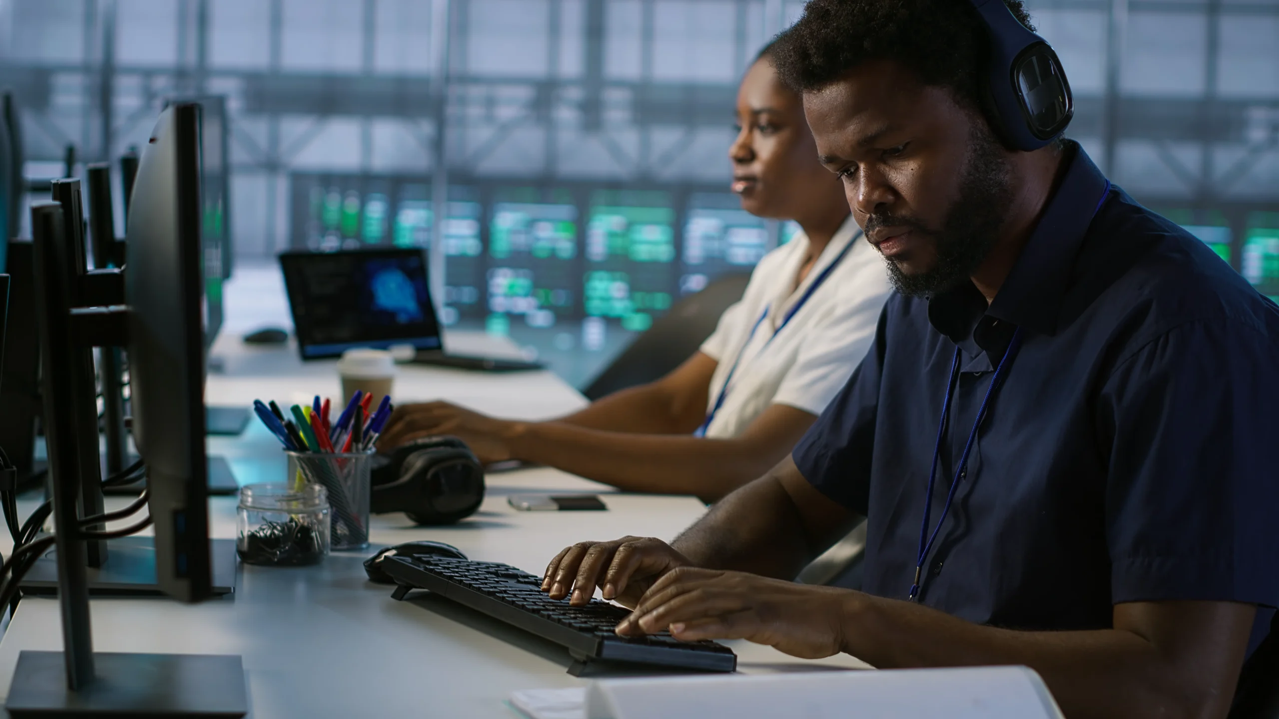 In a tech office, a focused man wearing headphones types on a keyboard. A woman works at a computer beside him. Multiple screens show data graphs 24/7 IT support at netsectechnologies